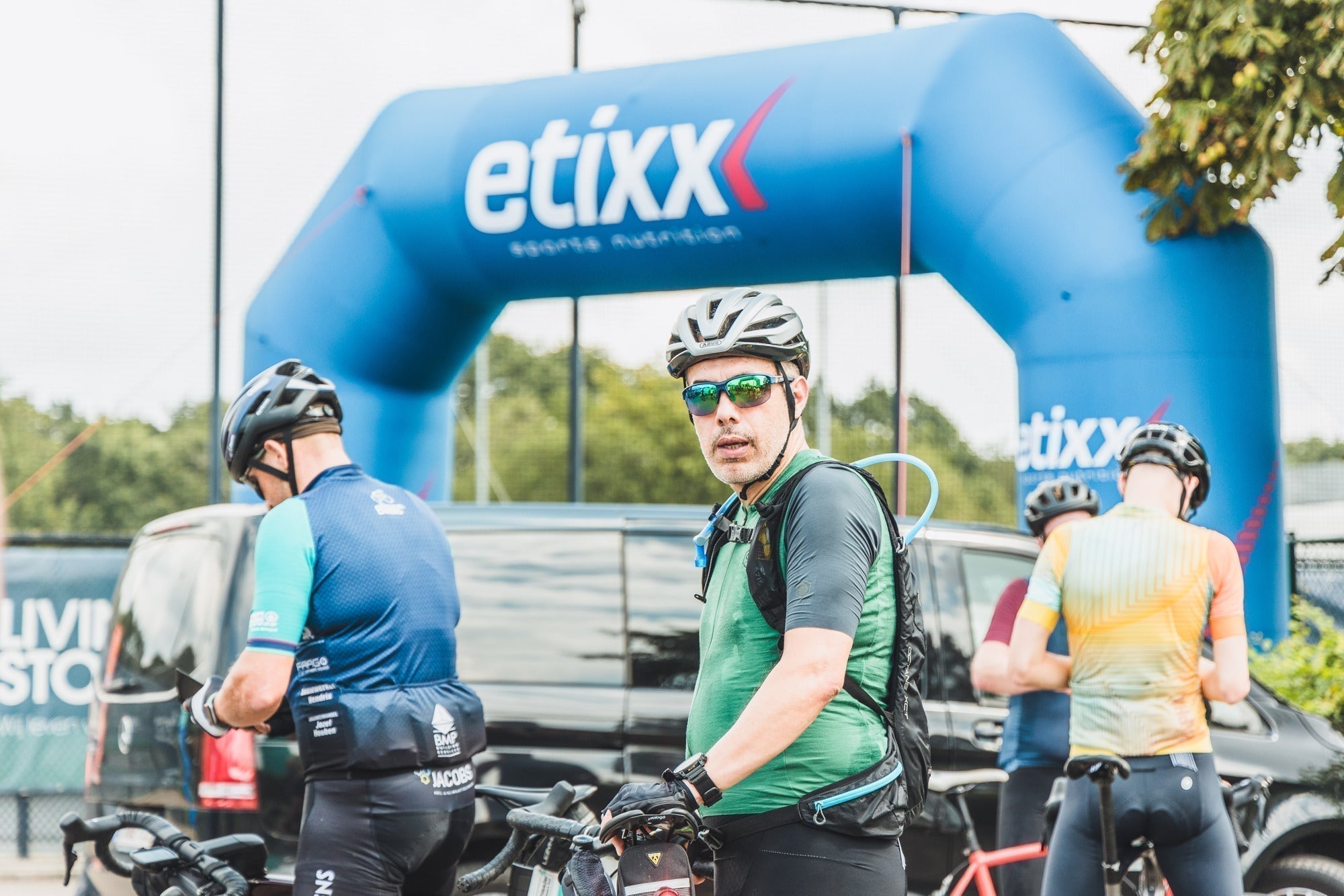 Cyclists standing together around the Scheldeprijs Cyclo event area, with one man in sunglasses looking into the camera
